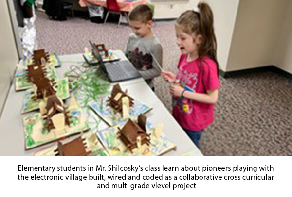 Two young students standing at a table looking  at a laptop and a cardboard pioneer village that the students built and connected to the laptop for light and audio display