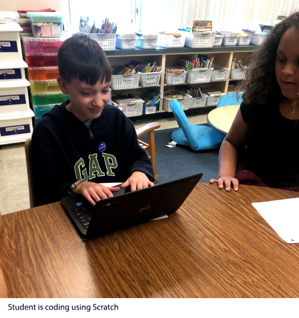 Student is sitting in front of a computer typing. He is learning to code using scratch. A girl sitting nearby looks on.